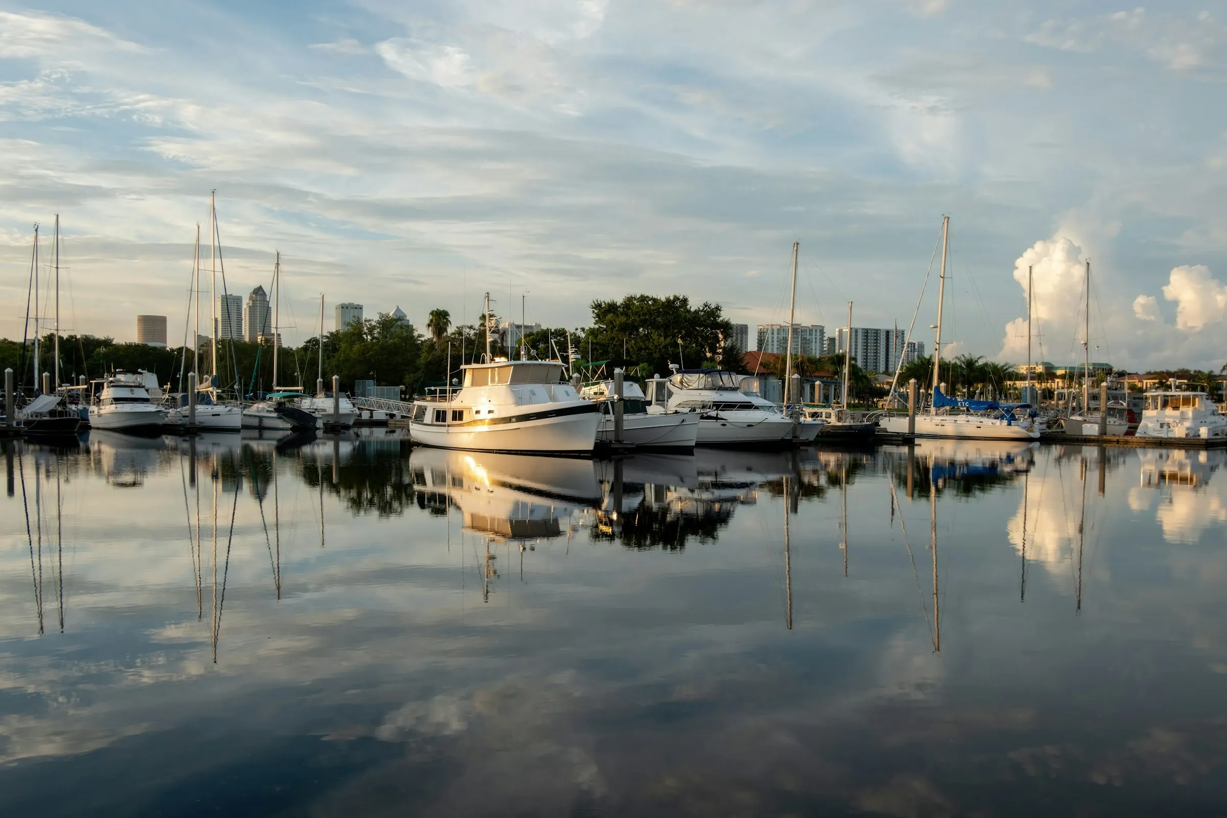Sailboats and a motor yacht mirrored in a calm harbor beneath a distant skyline