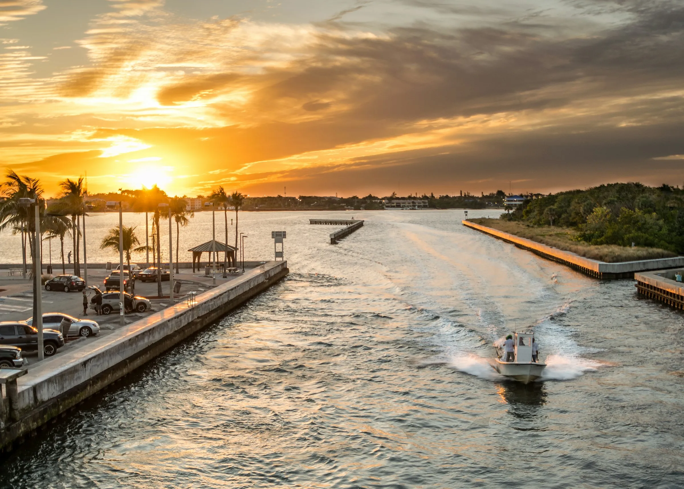 A small powerboat heading out an inlet past a palm-lined seawall at sunset