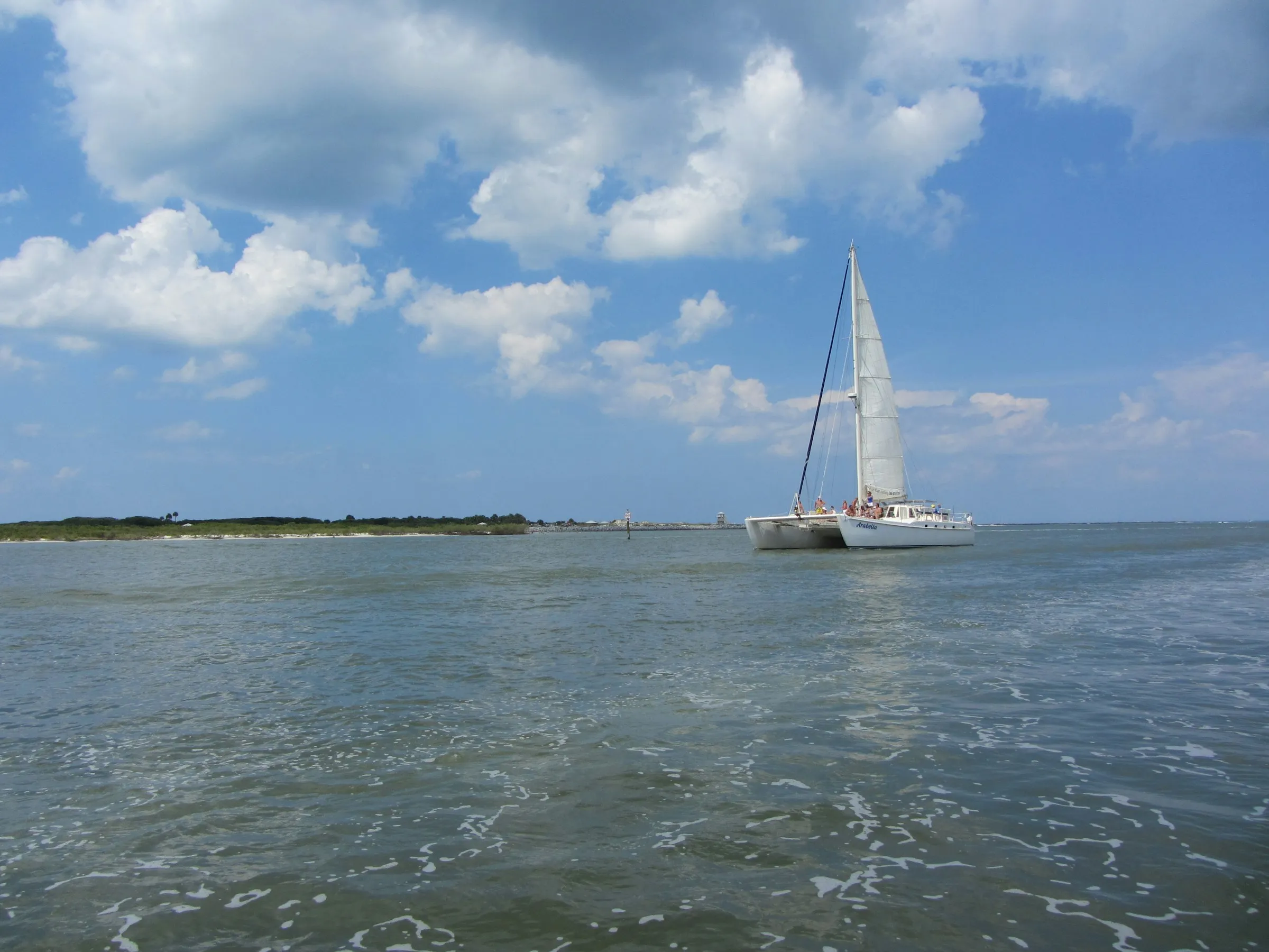 A catamaran under sail off a low barrier island under towering clouds