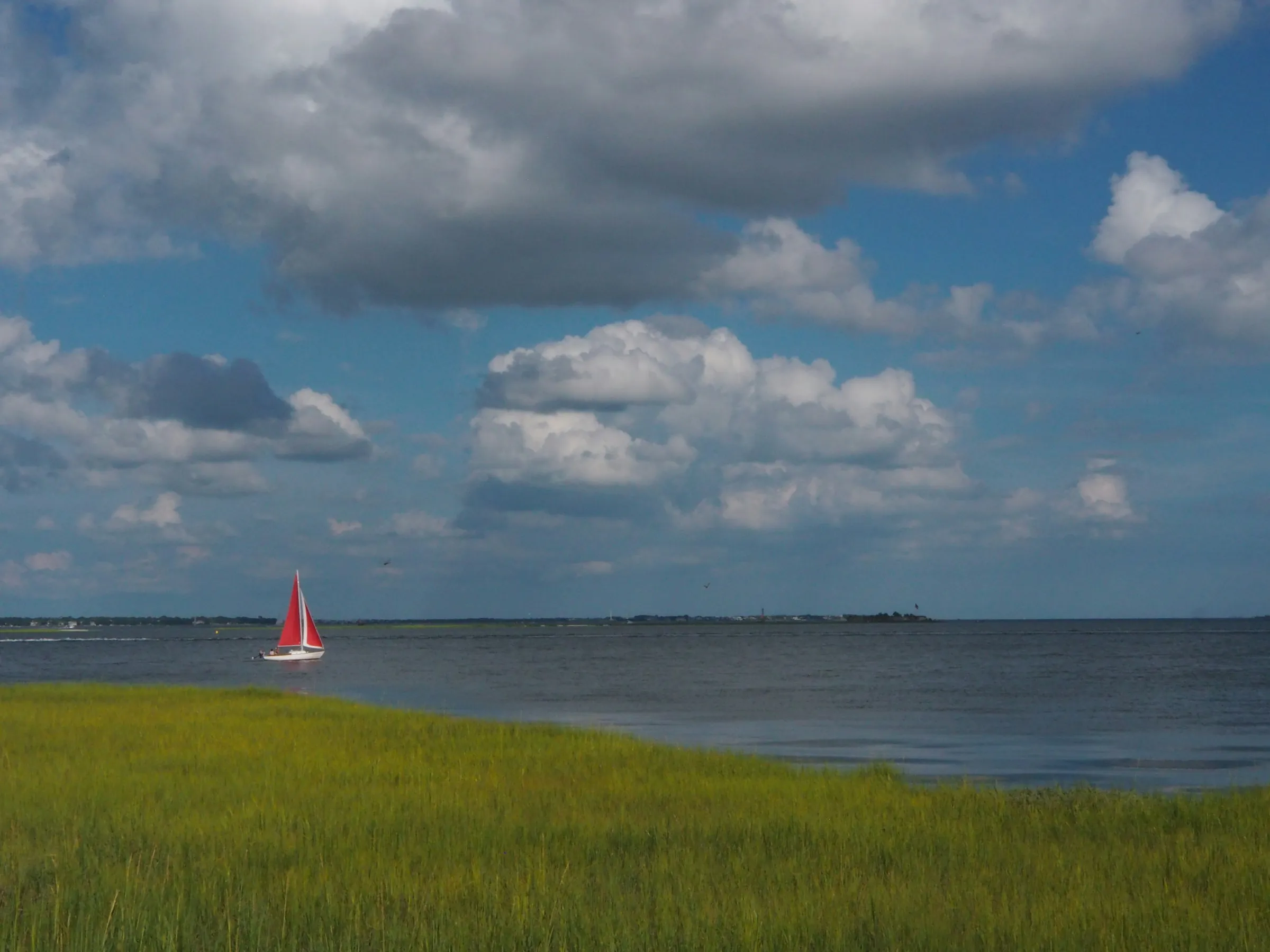 A red-sailed boat on open water beyond a green salt marsh under tall cumulus cloud