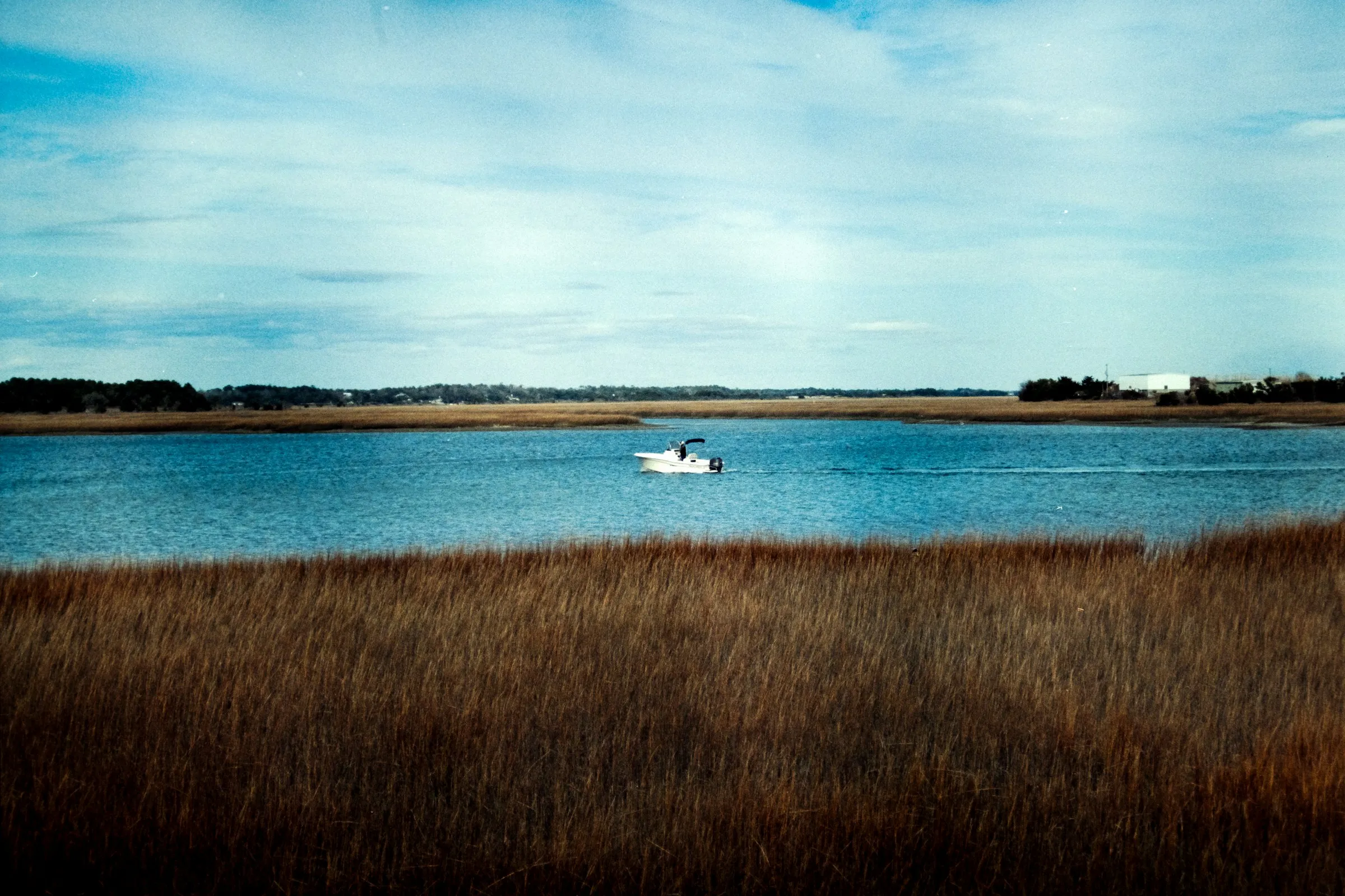 A small boat crossing a wide tidal river seen over golden marsh grass